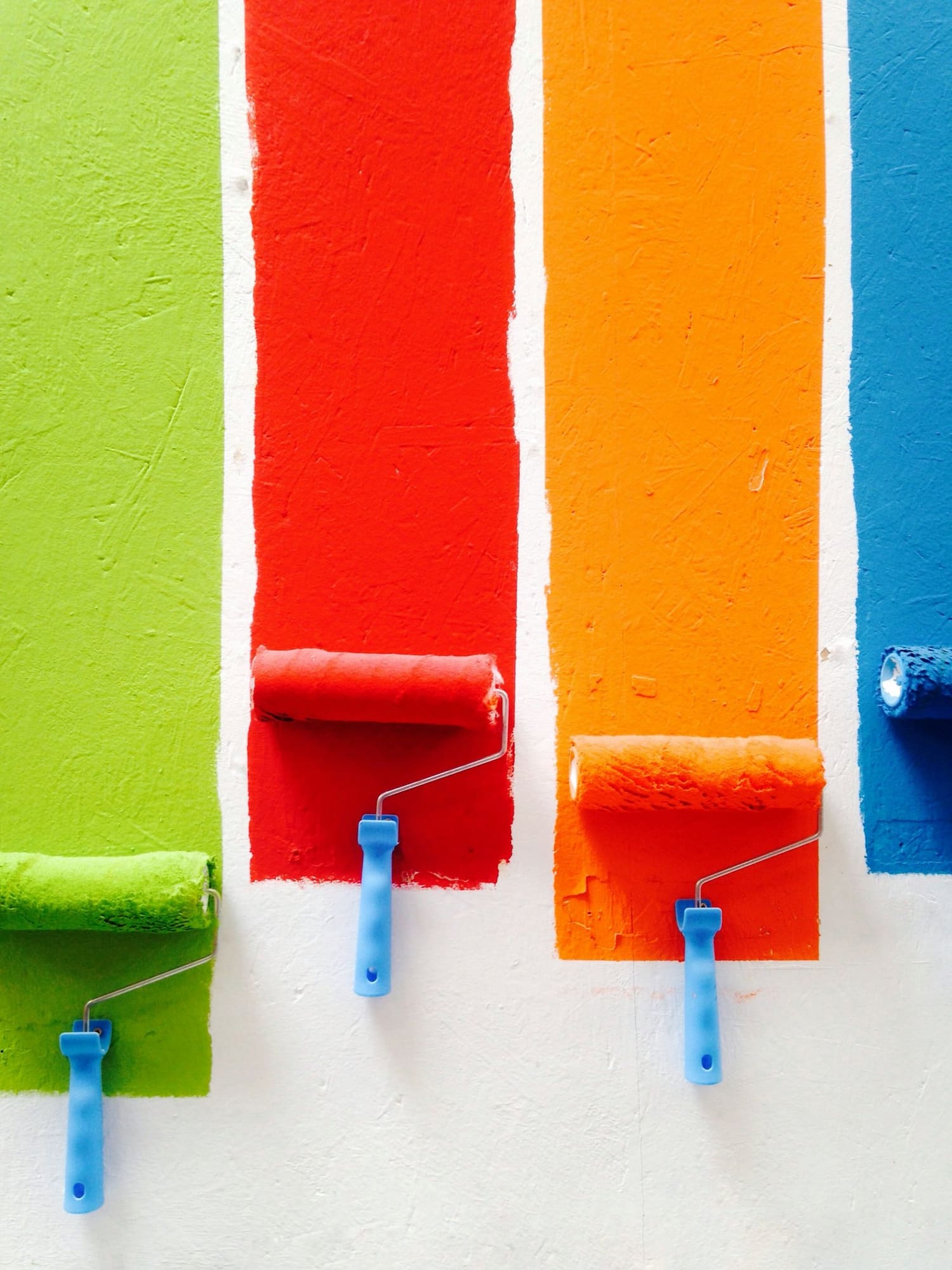 Paint rollers applying bright green, red, orange, and blue paint to white wall stripes
